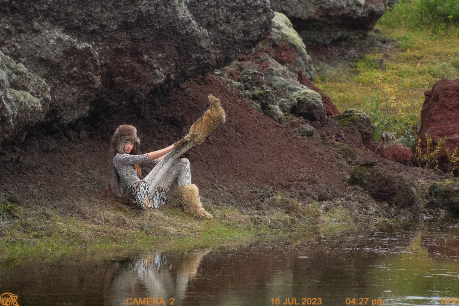 Fashion model in brown fur-like sleeves and patterned trousers reclining against rocks beside a pond.