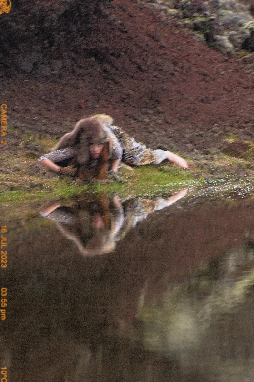 Person in textured animal-print clothing lying beside a pool of water surrounded by dark reddish earth.
