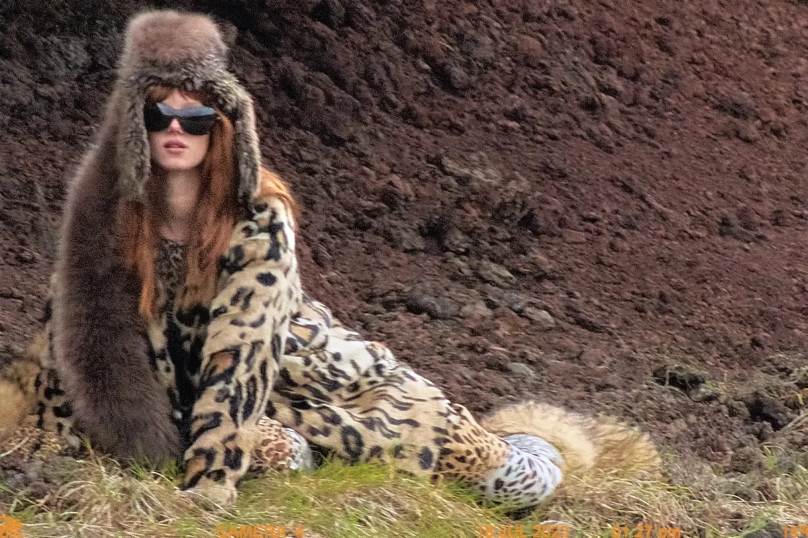 Fashion portrait of a red-haired model in oversized brown furry headwear, dark sunglasses and animal-print outerwear.