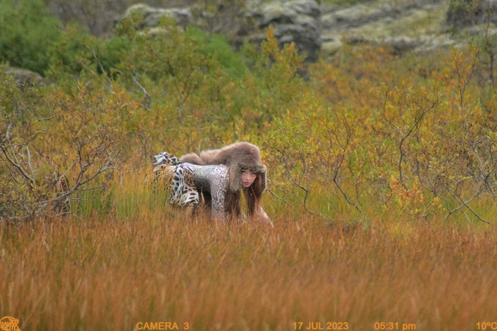 Person wearing textured animal-print clothing bending in a grassy Icelandic field.