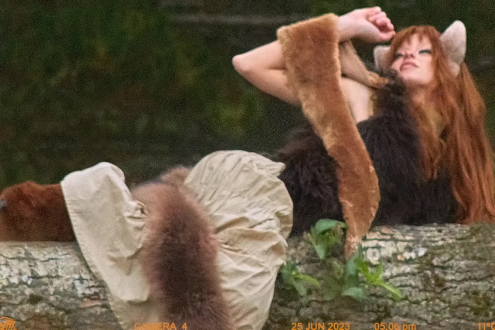 Red-haired model wearing long brown furry sleeves reclining on a stone ledge by dark green water.
