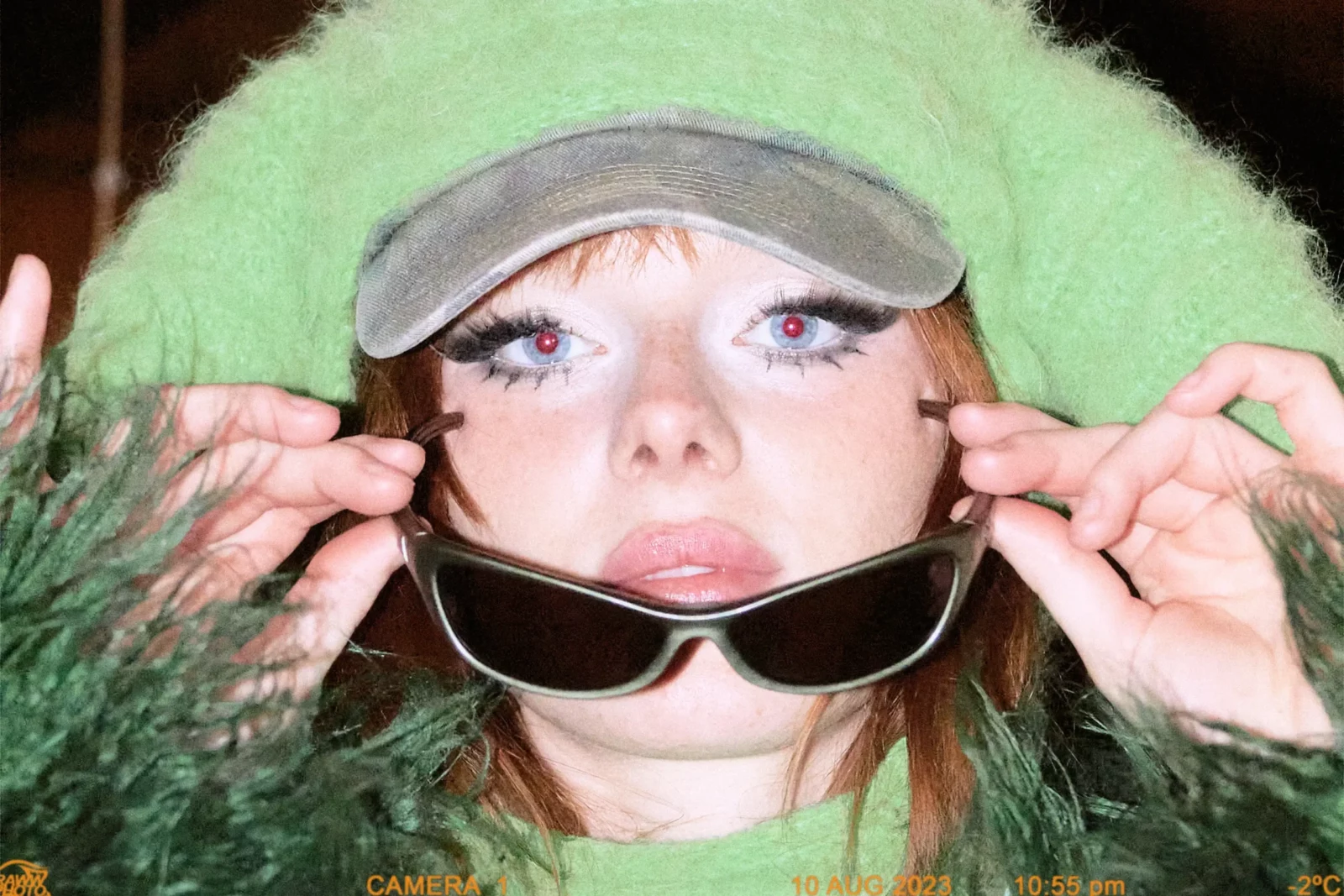 Close-up portrait of a woman in fluffy mint-green headwear adjusting narrow black sunglasses.