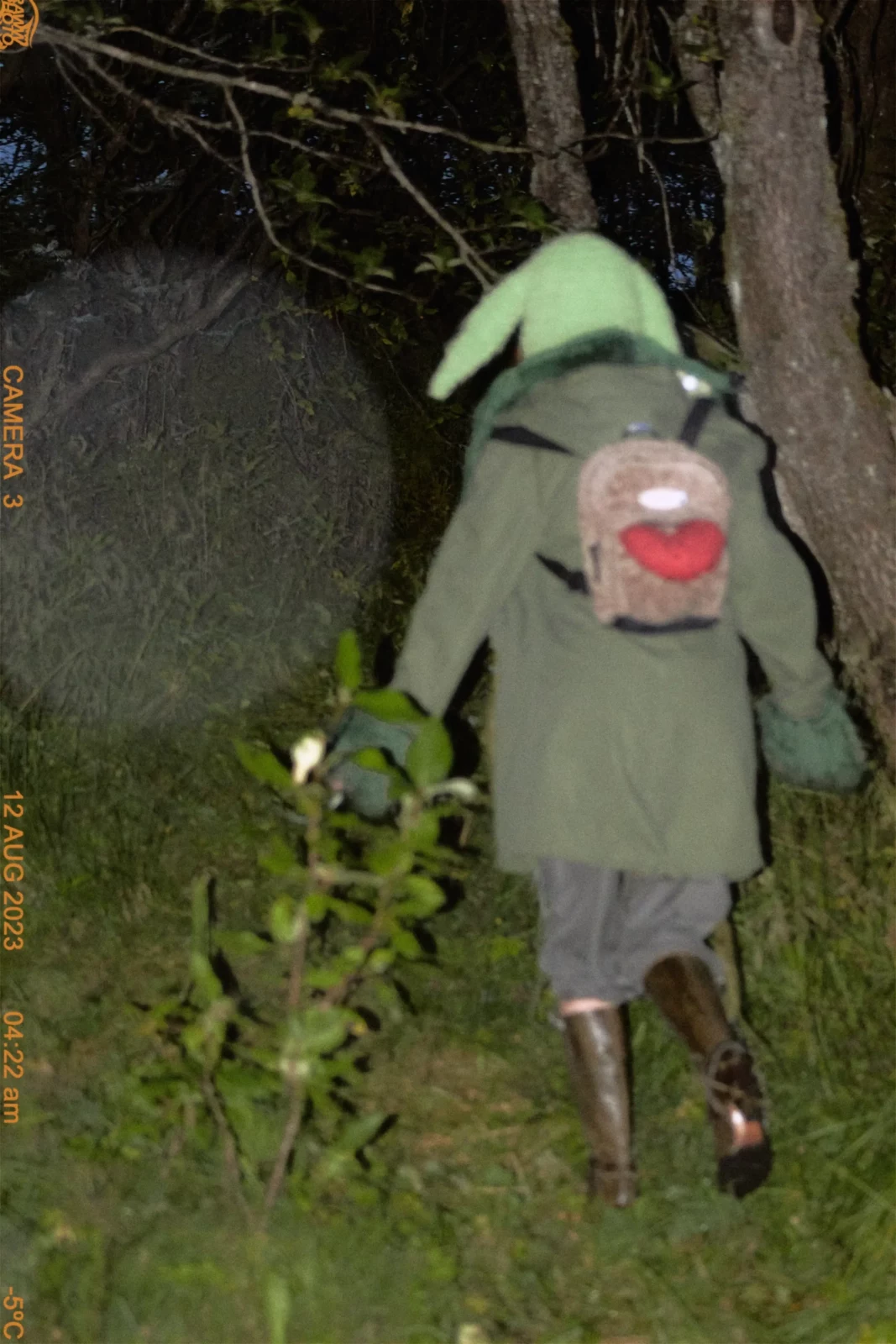 Rear view of a model in a pale green raincoat and backpack walking into a wooded area.