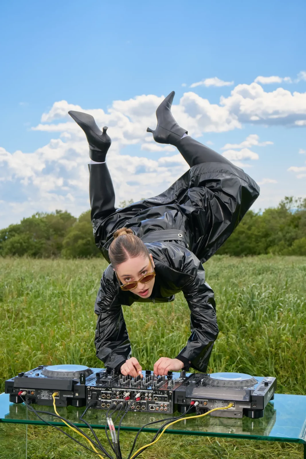 Model in dark clothing balancing upside down in a grassy field under a cloudy blue sky.