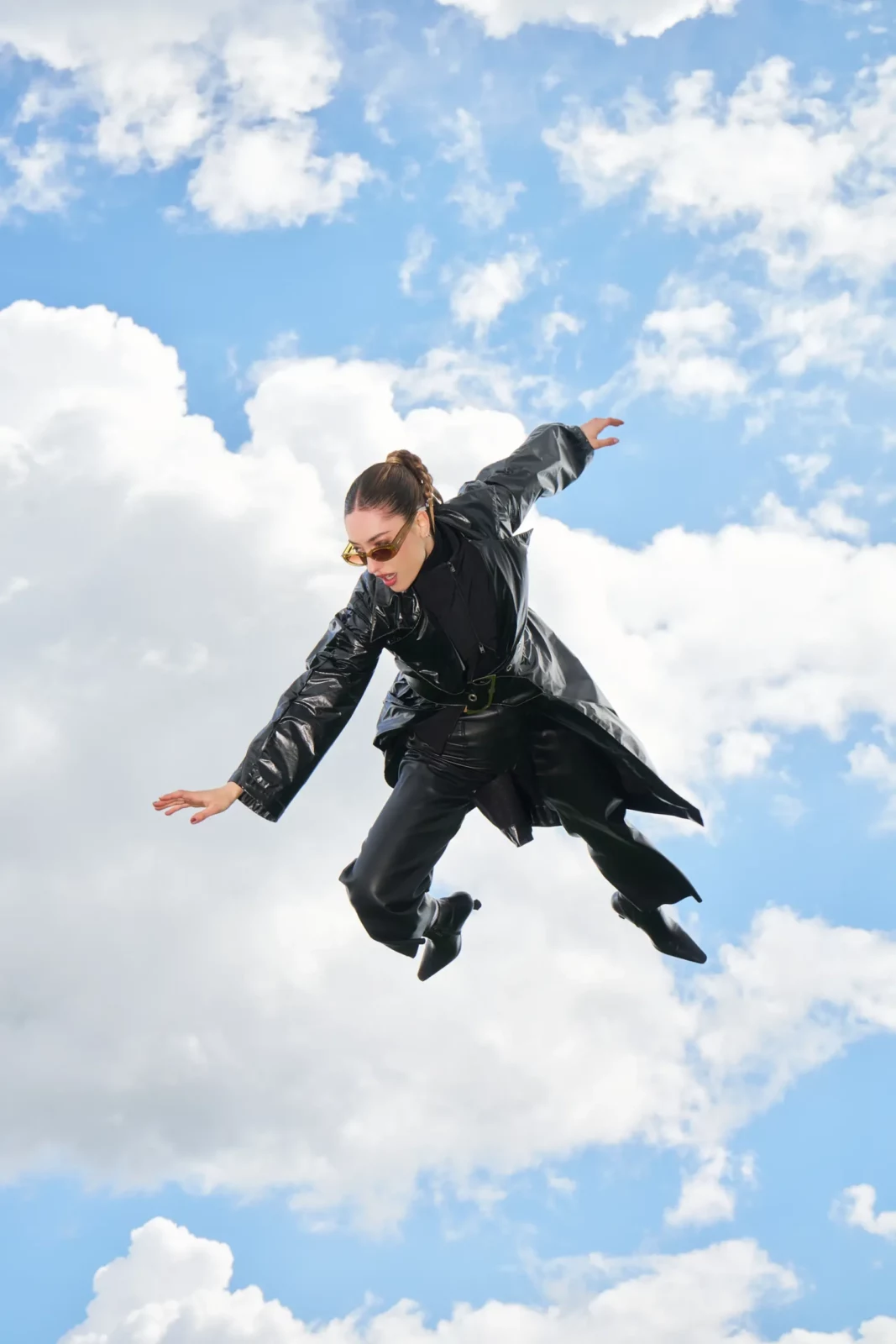 Model in a black outfit leaping against a bright blue sky with scattered clouds.