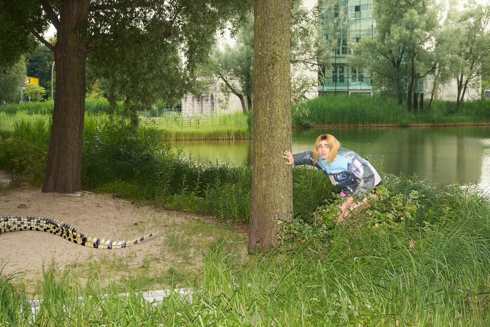 Model in a colorful patterned outfit crouching near a lakeside tree in a green Berlin park.