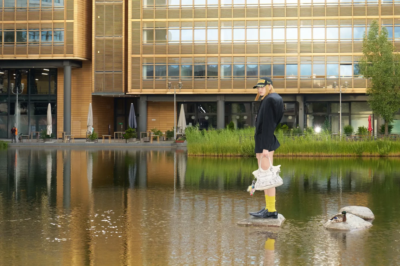 Fashion model in a black jacket and yellow socks standing beside a pond with a swan.
