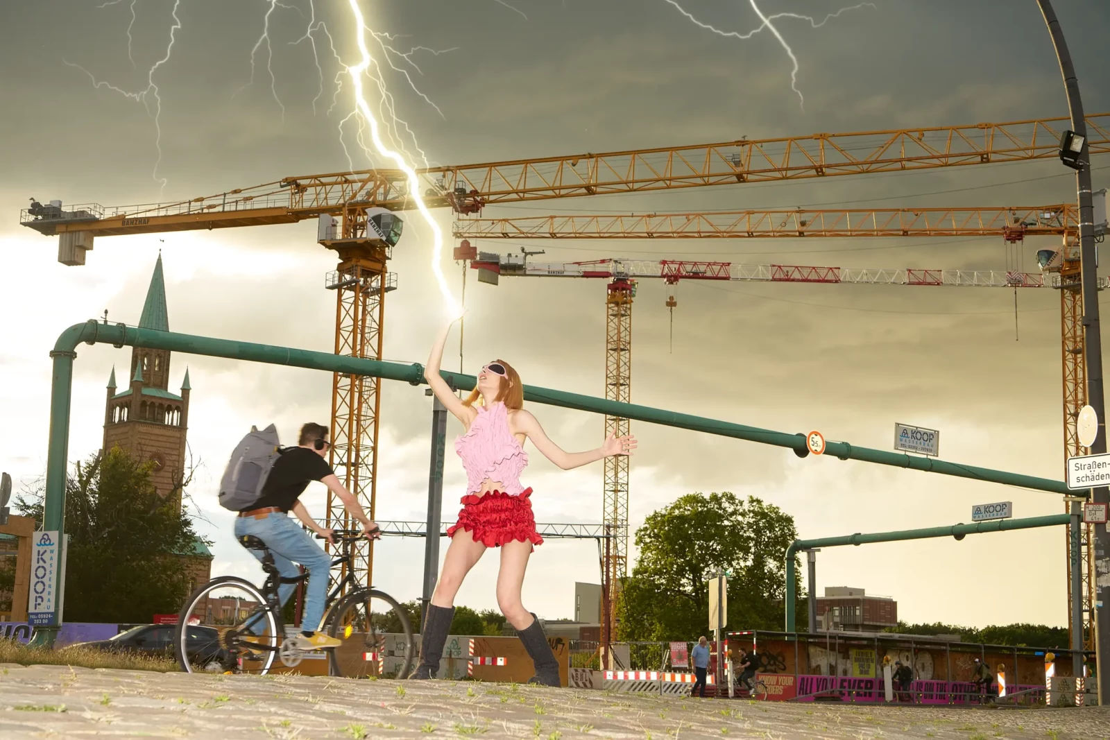 Model wearing a pink ruffled skirt posing near cranes and a cyclist beneath a lightning-filled sky.