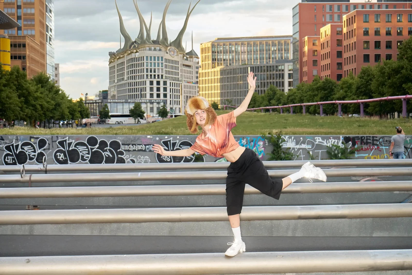 Fashion model in a pink top and black shorts balancing on a metal barrier beside Berlin city traffic.