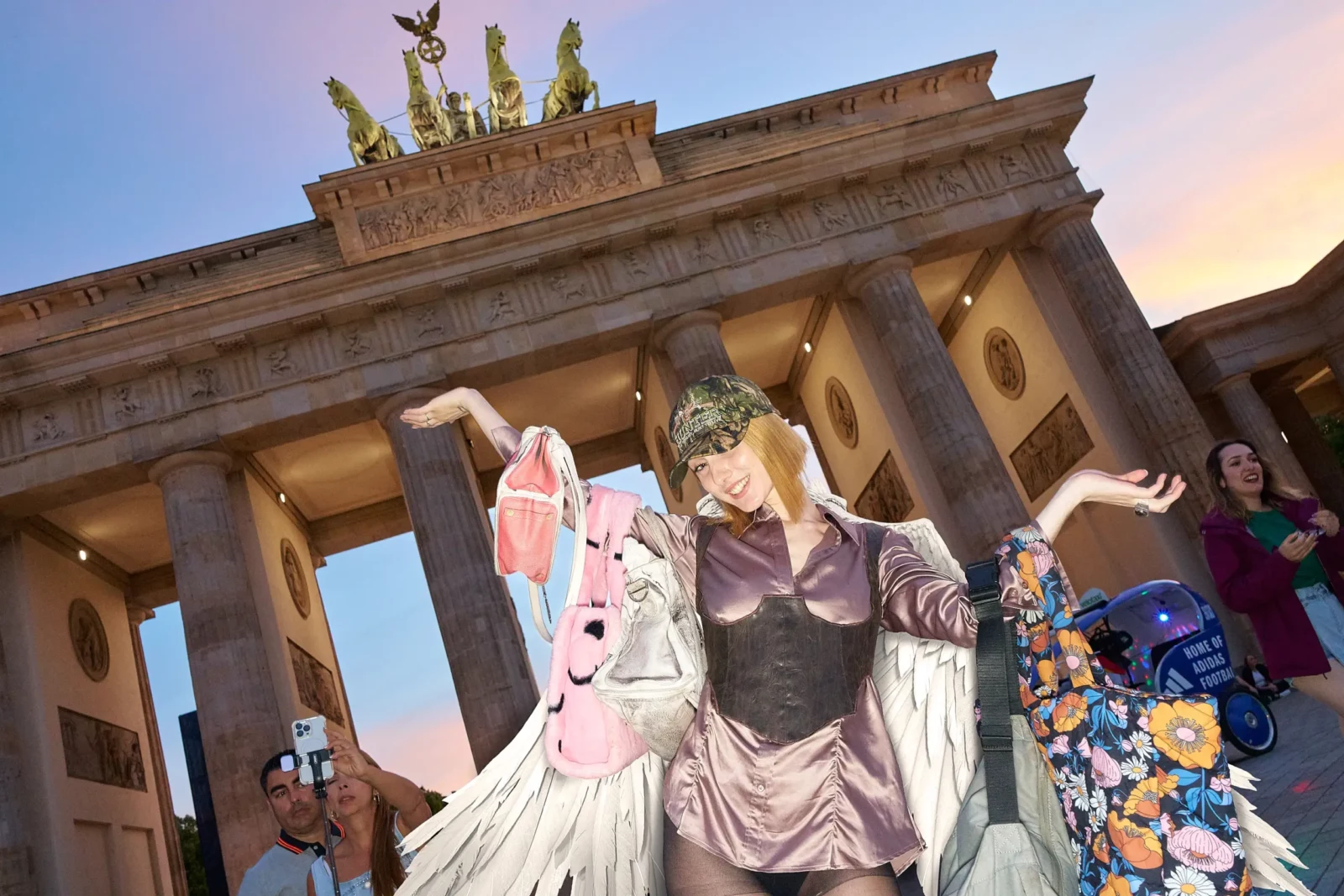 Avant-garde fashion model in a sculptural outfit holding colorful accessories in front of Berlin's Brandenburg Gate at sunset.
