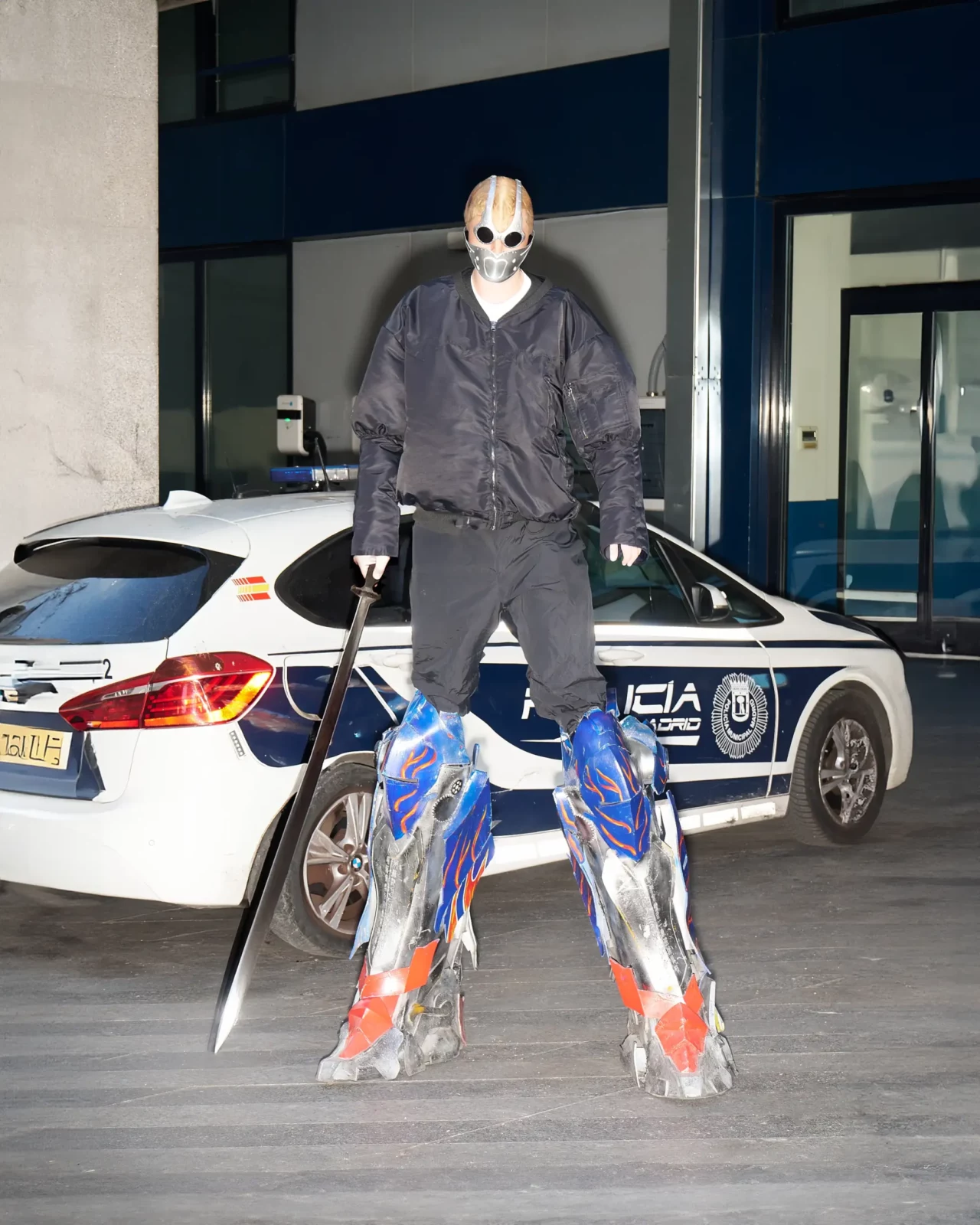 Masked figure in a dark outfit standing beside a parked white car and holding bright multicolored fabric sculptures.
