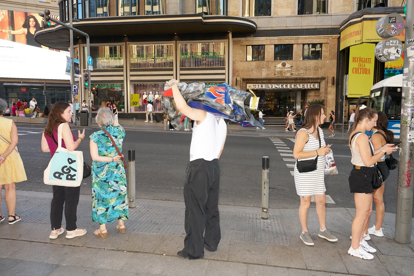 Outdoor street scene with several people in colorful outfits standing and gesturing near a bus shelter.