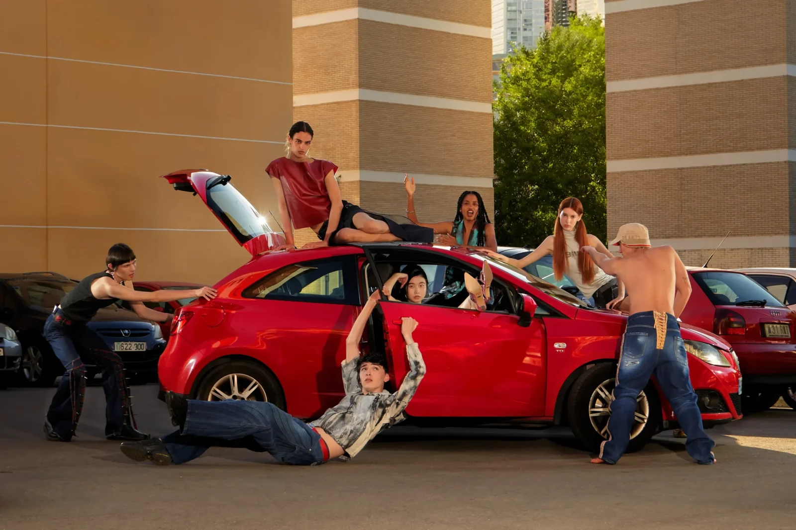 Group of fashion models gathering around a red car in an urban parking area.