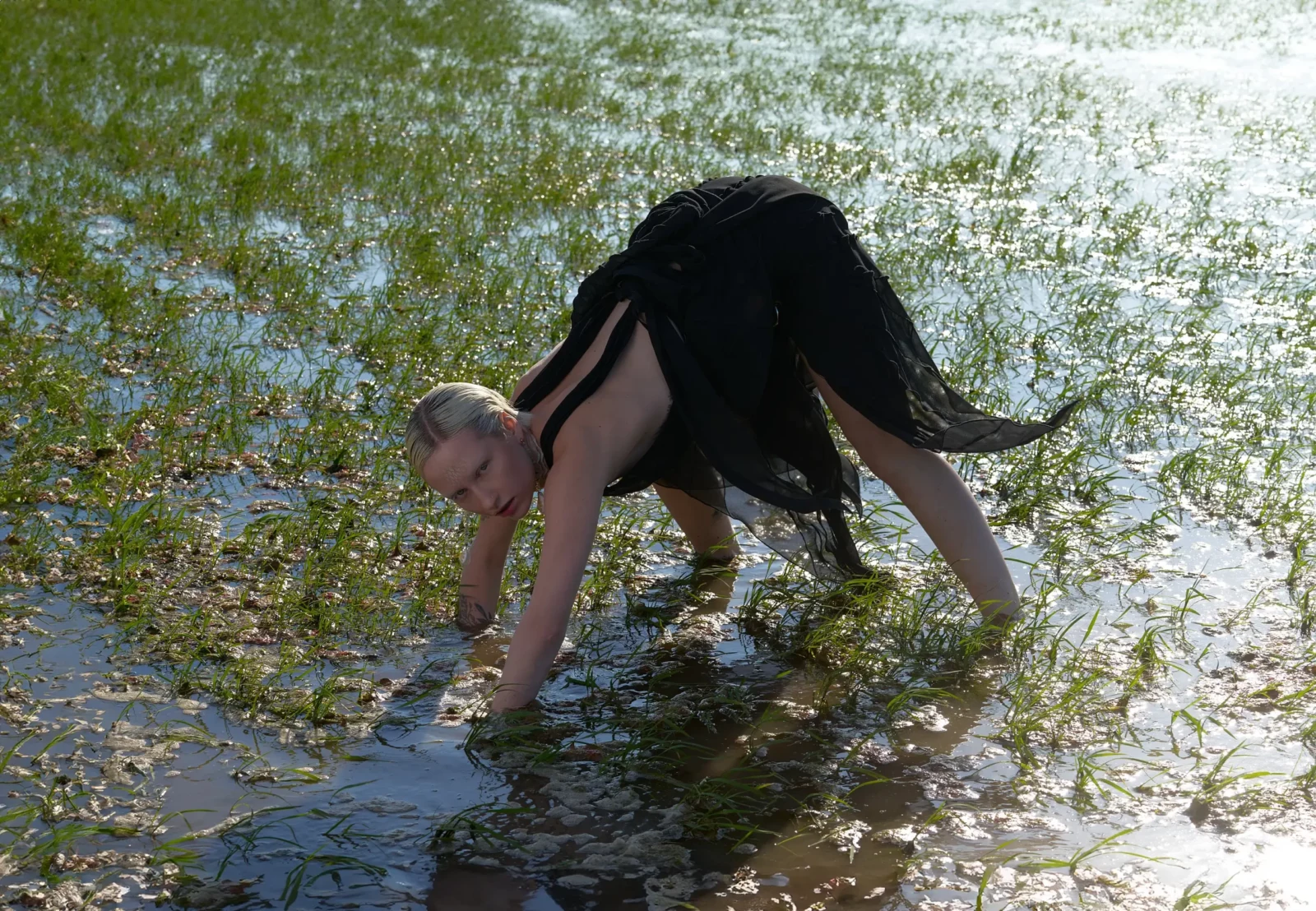 Fashion model in a black outfit crawling through wetland grass and reflective puddles.