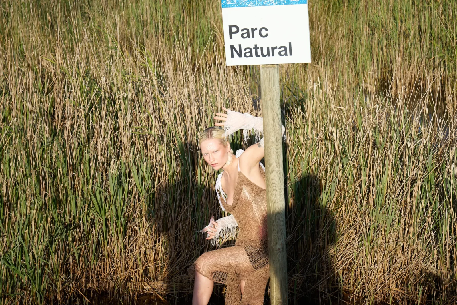 Model wearing a mesh-textured outfit posing beneath a nature reserve sign among reeds.