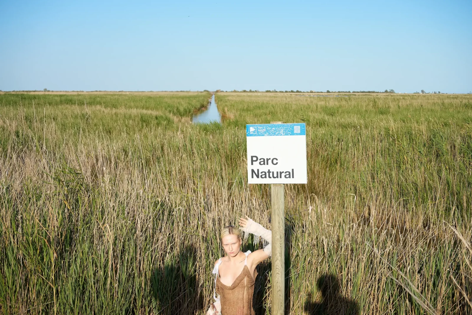 Distant fashion model crouching in tall grasses below a nature reserve sign.