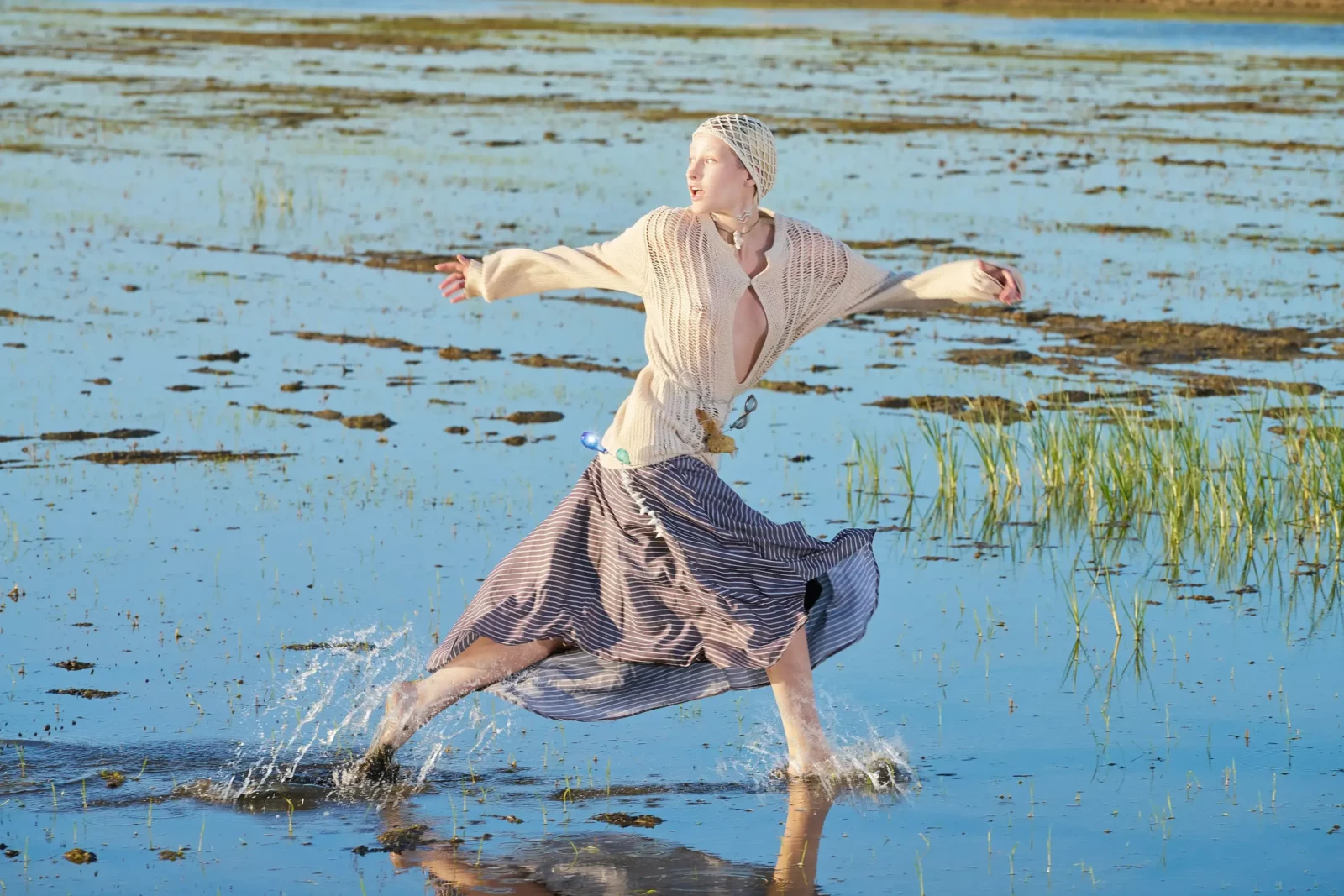 Blonde fashion model moving barefoot through shallow water in a flowing outfit.