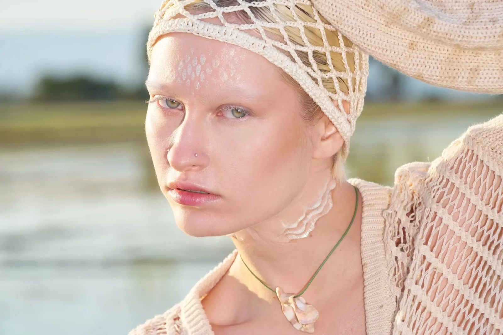 Close-up of a blonde model wearing a mesh headpiece and layered jewelry by the water.