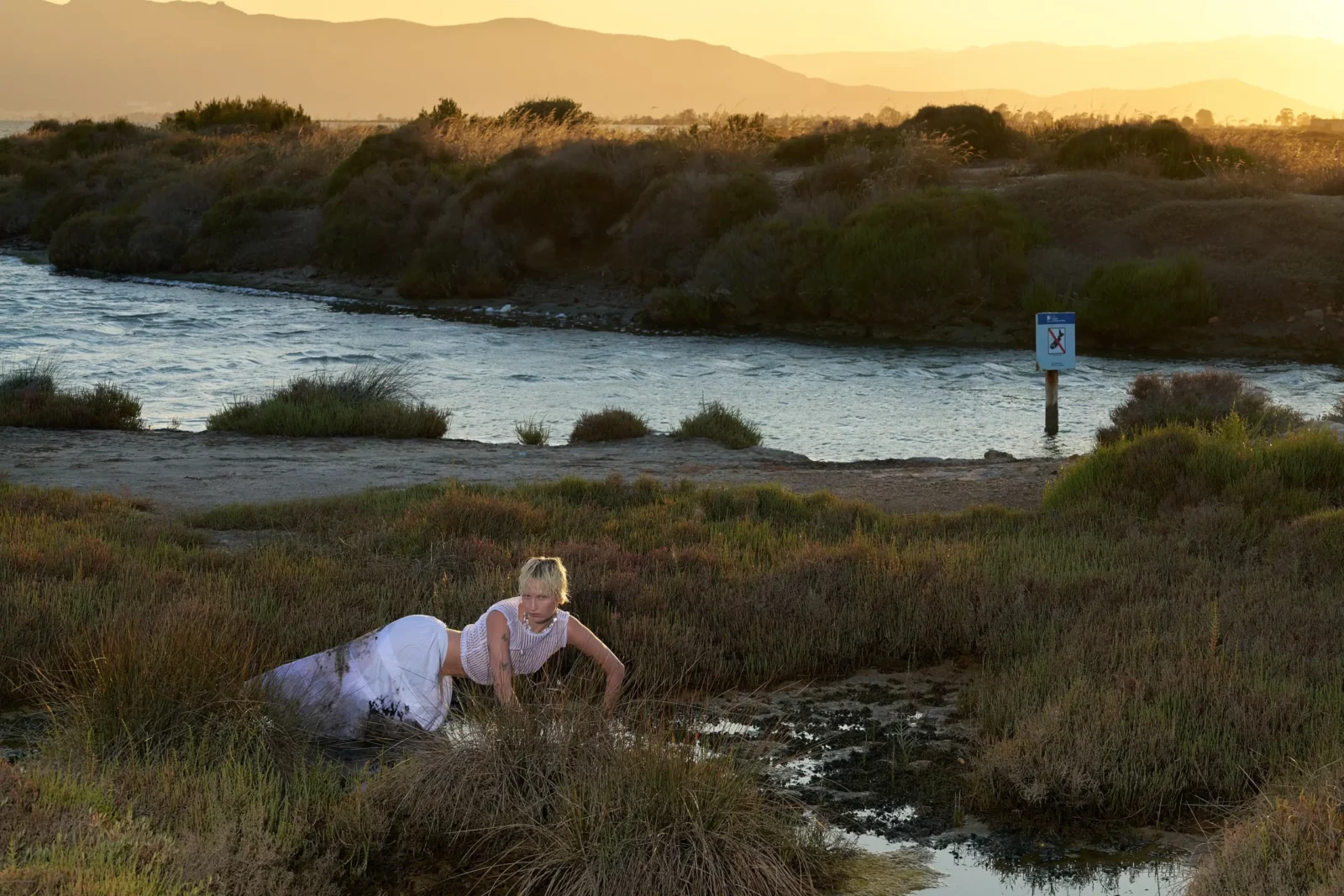 Model reclining in marsh vegetation beside a still lagoon at golden hour.