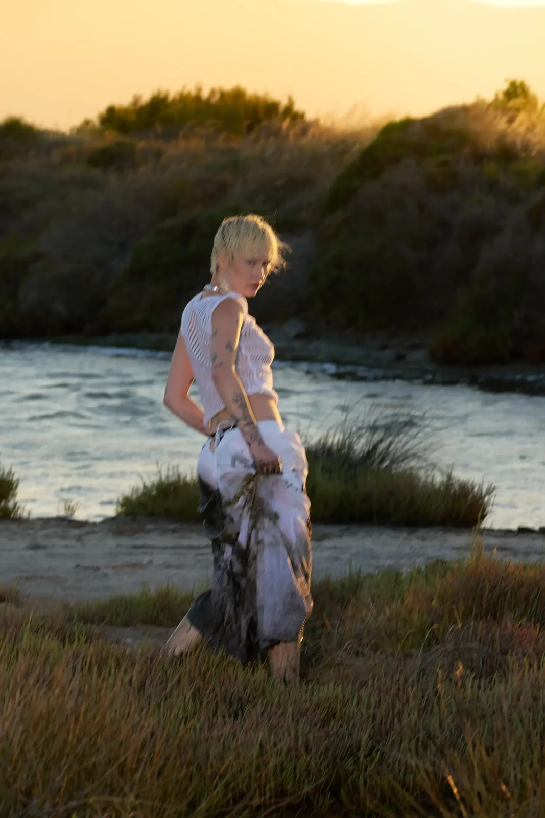 Blonde fashion model in a pale outfit walking through marsh grass near the water.