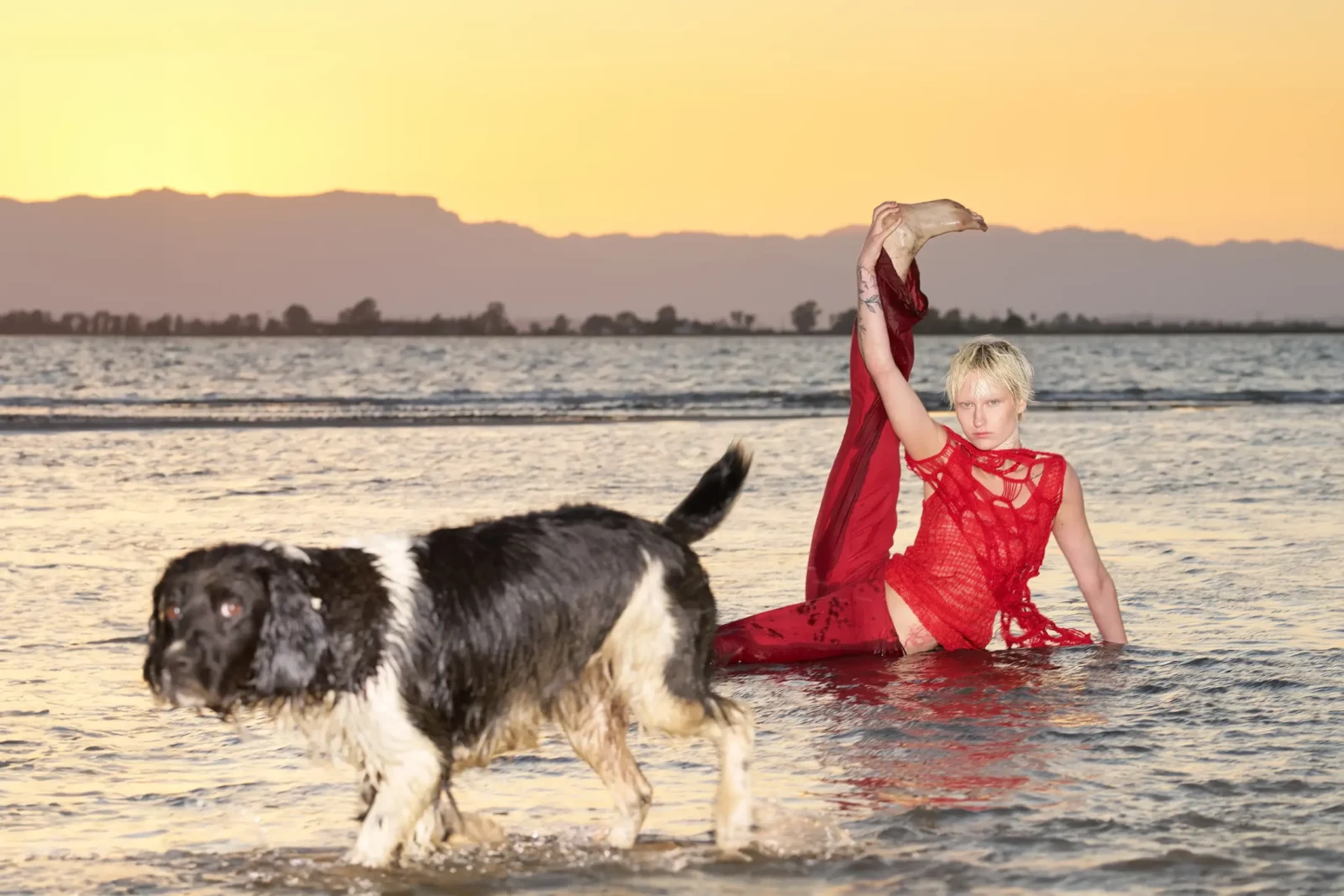 Model wearing vivid red clothing and raising an arm beside a dog in the water at sunset.