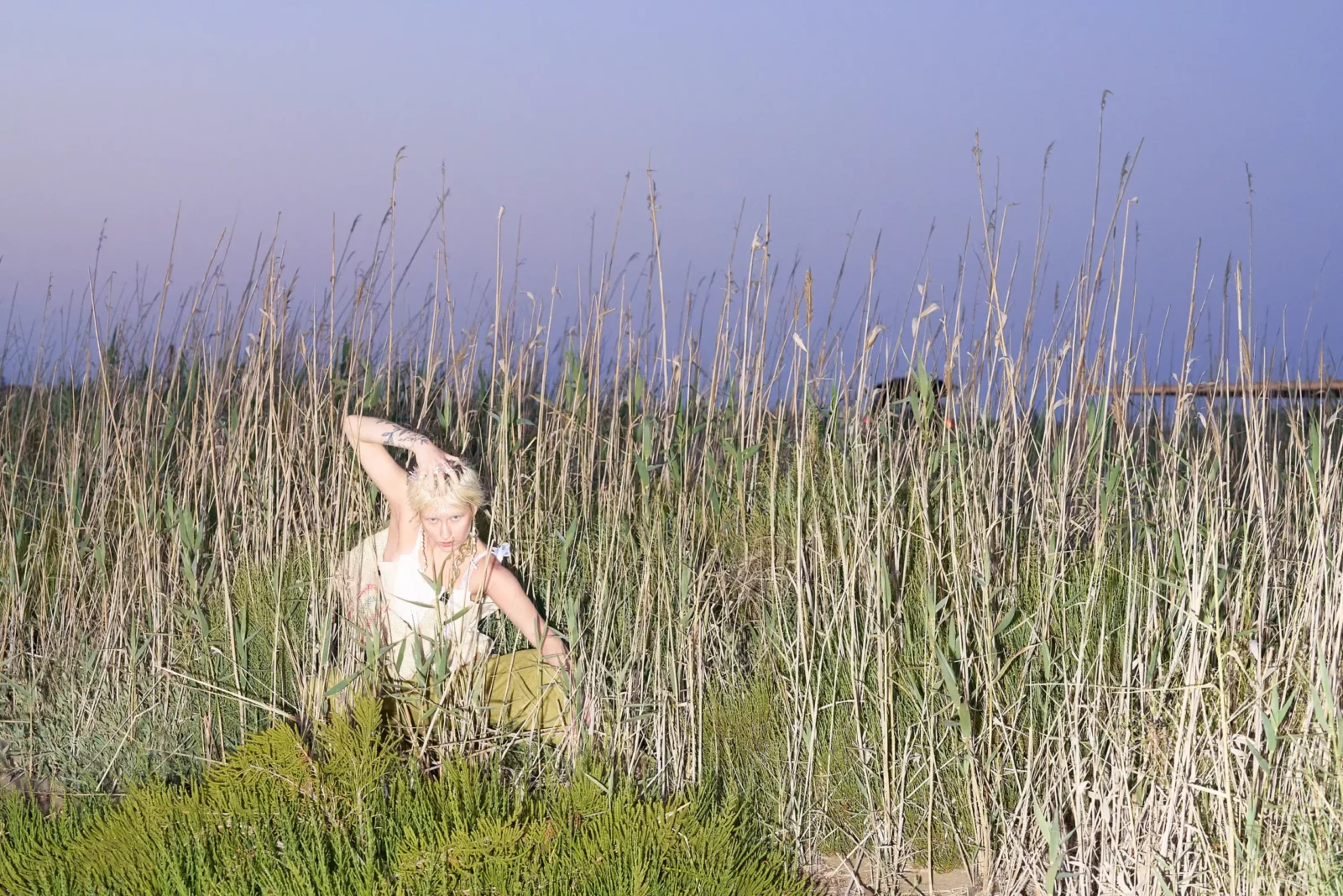 Model in a pale outfit partly concealed by tall reeds against a lavender evening sky.