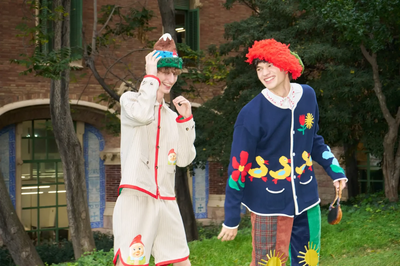 Two fashion models outdoors in playful oversized outfits, one in a cream set and the other with a bright red fluffy hat.