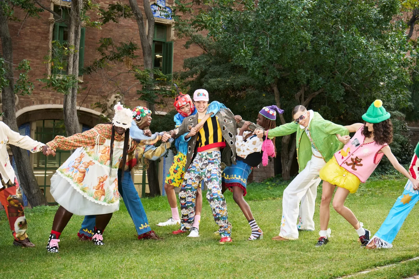 Group of fashion models in vivid multicolored outfits holding hands in a circle on a grassy urban lawn.