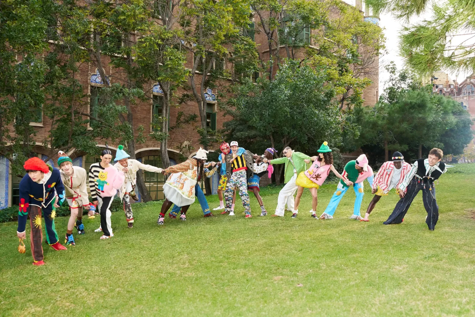 Wide fashion campaign photograph of a large group in colorful patterned clothing forming a chain across a park lawn.