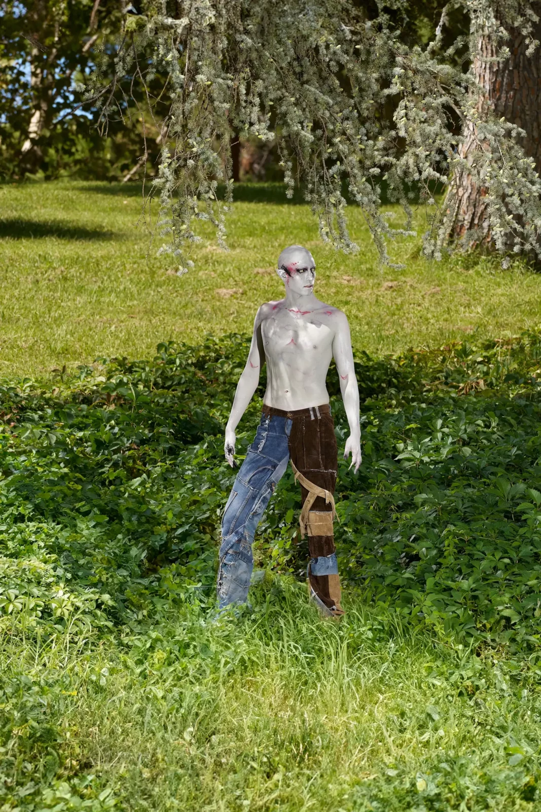 Silver-faced figure standing alone in tall grass under leafy trees.