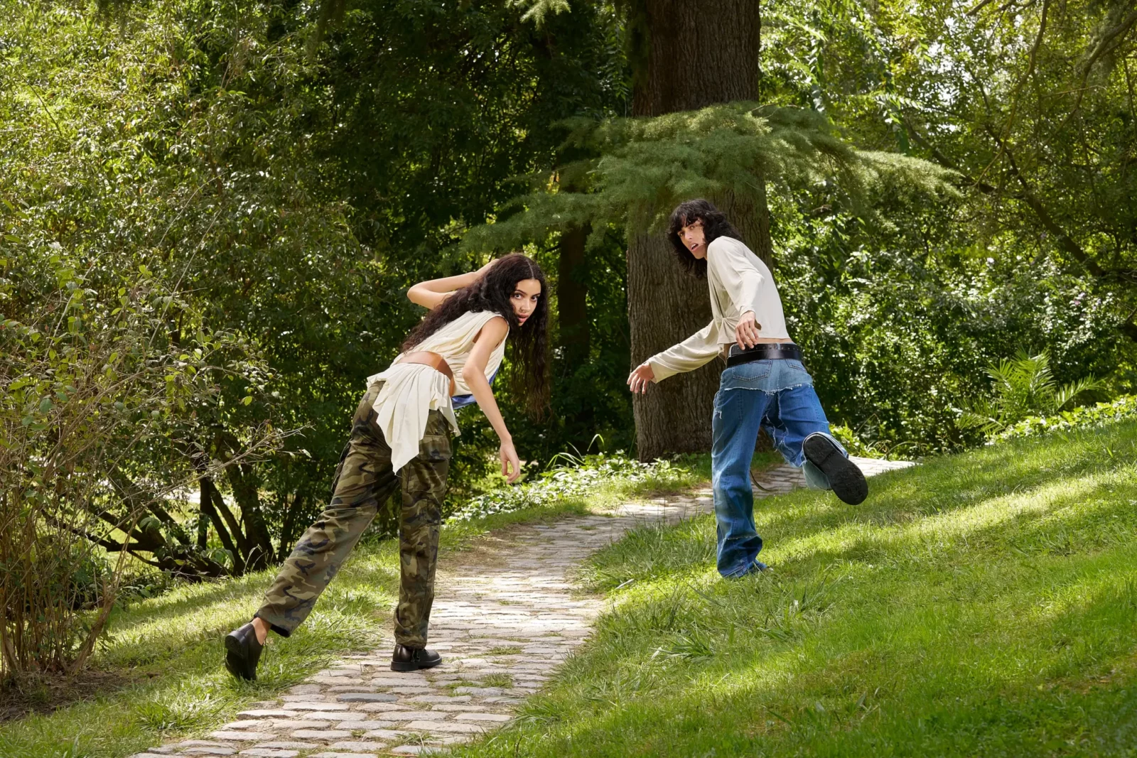 Two fashion models moving along a park pathway bordered by green plants.