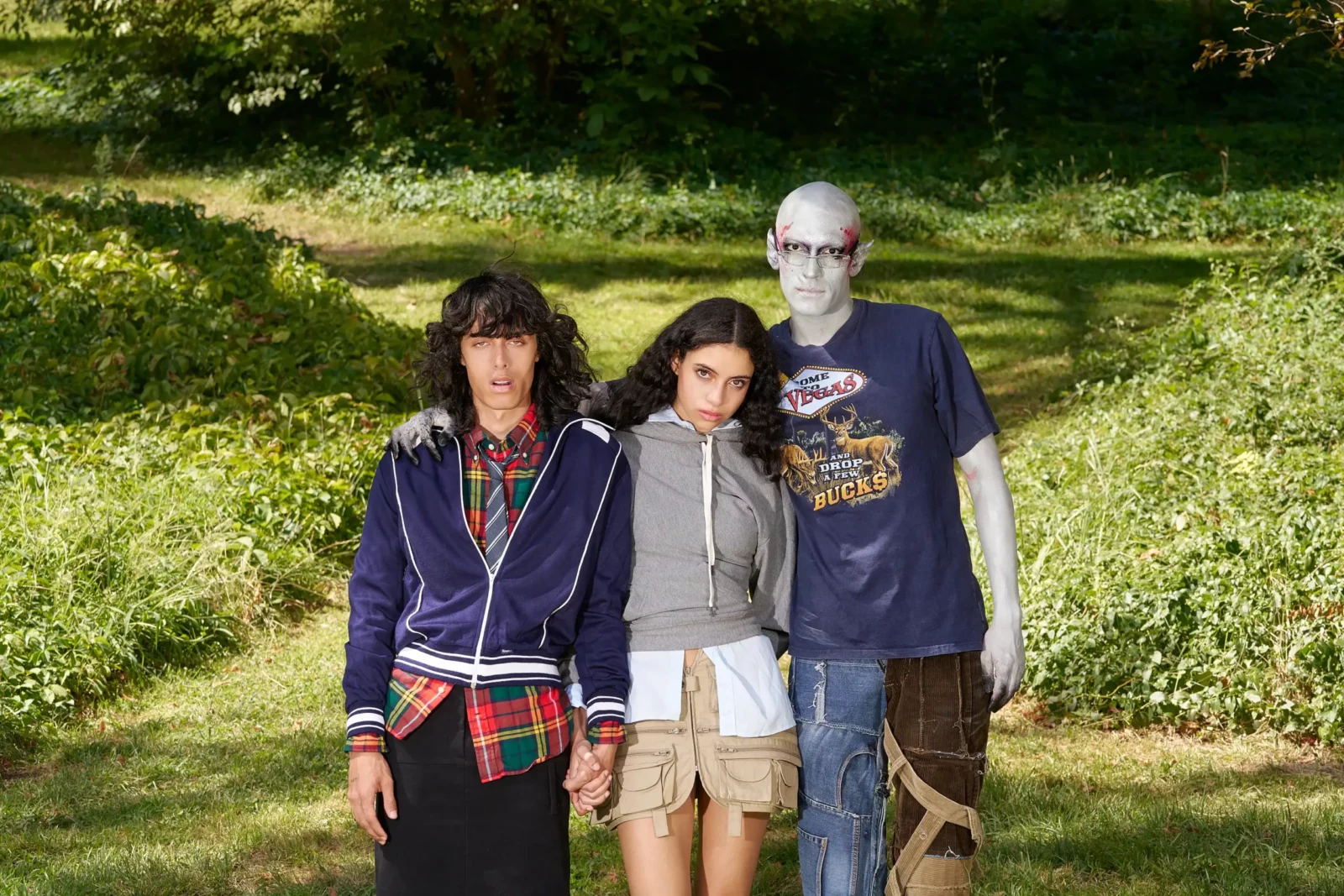 Three fashion characters, including a silver-faced figure, posing together in a sunlit park.