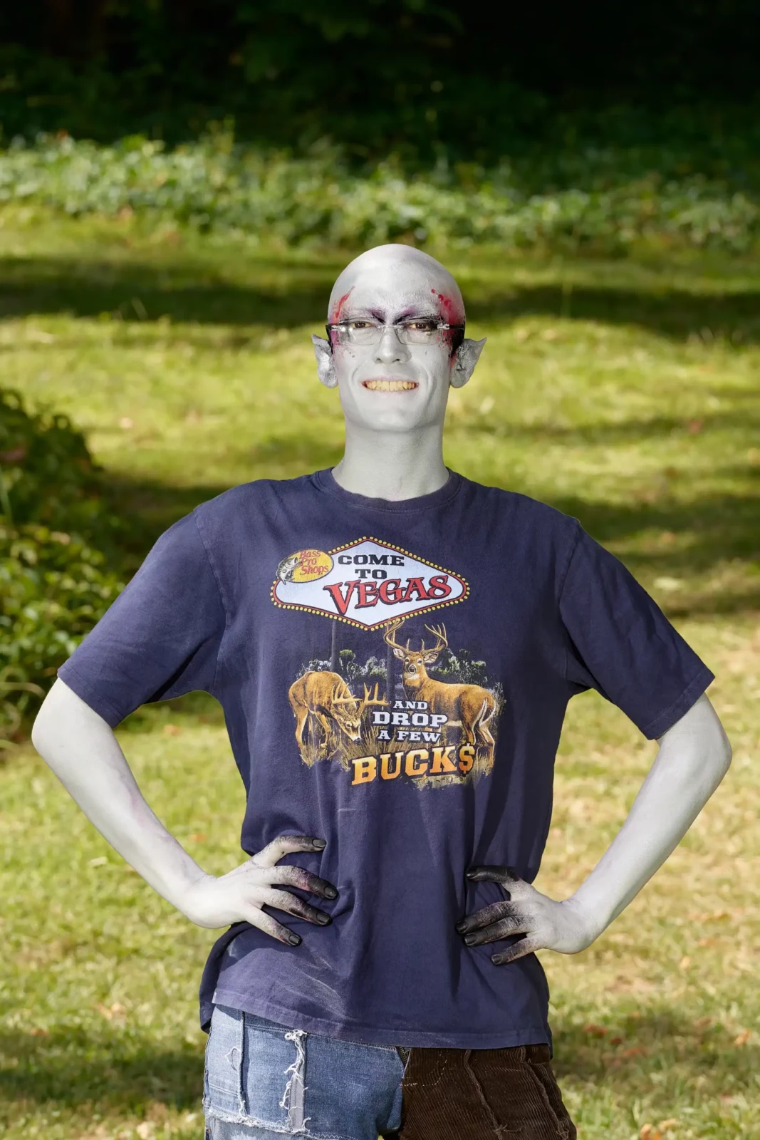 Silver-faced alien character in a graphic T-shirt posing with hands on hips in a park.