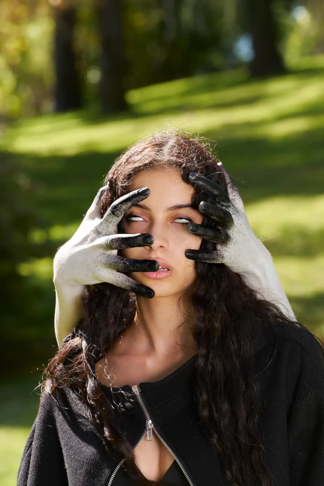 Dark-haired model wearing black gloves framing her face outdoors among trees.