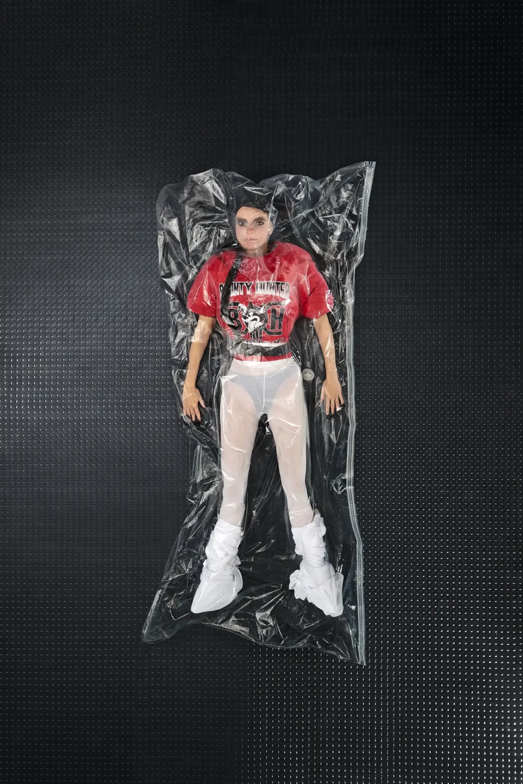 Overhead fashion photograph of a model in a red jersey and white trousers inside a clear plastic sheet.