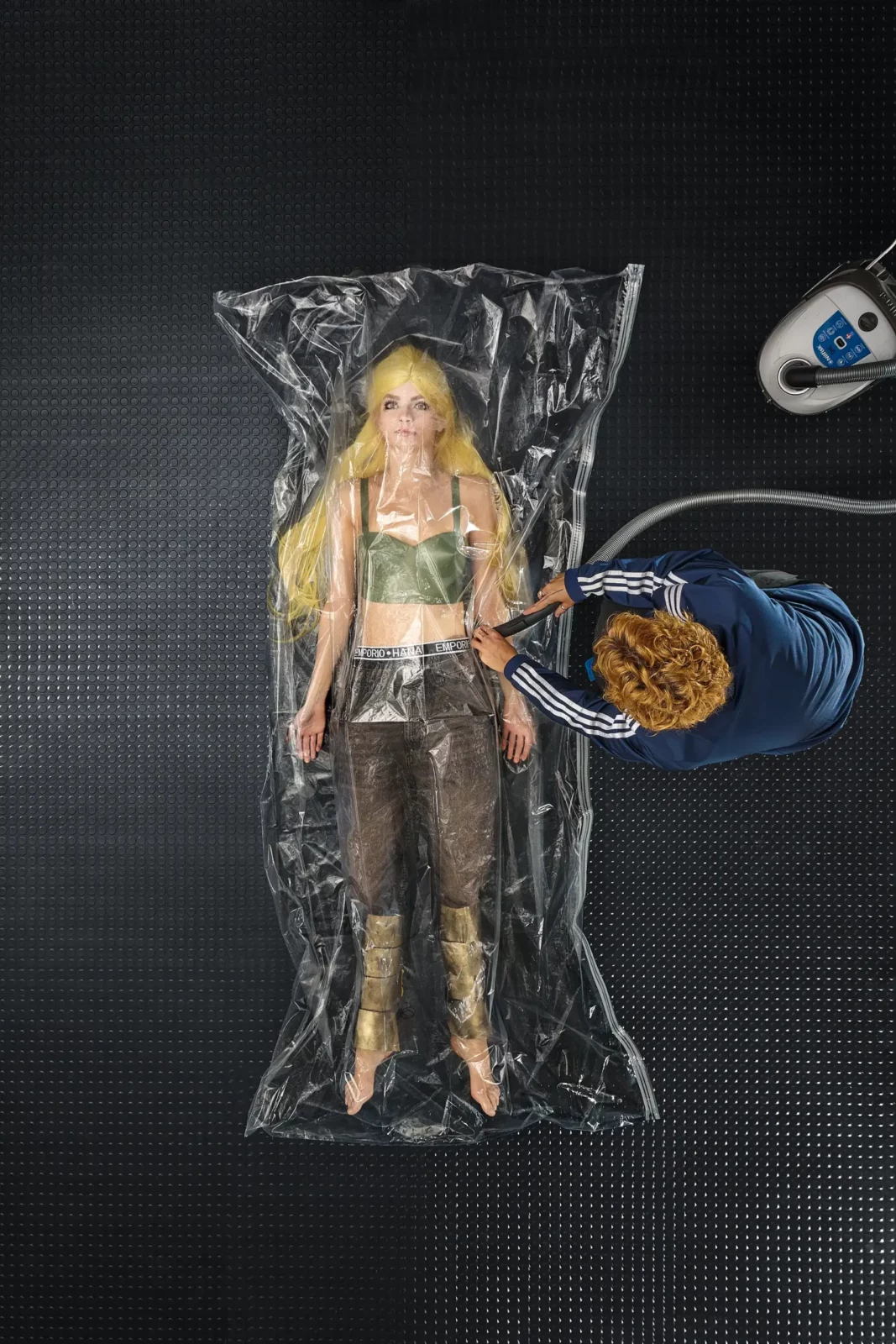 Overhead fashion photograph of a blonde model in metallic green clothing lying inside transparent plastic sheeting.