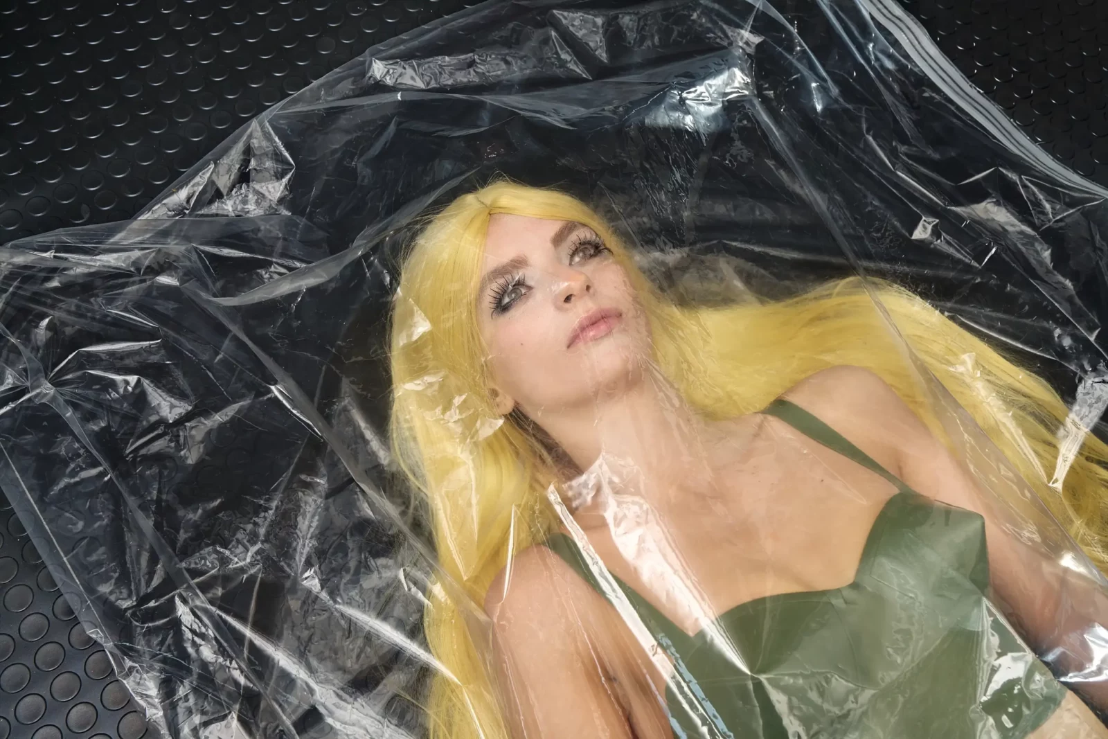 Close-up of a blonde model in a green bustier lying beneath crinkled transparent plastic.