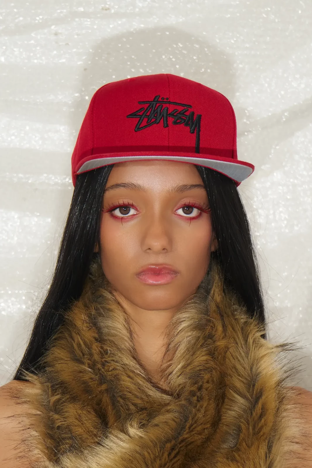 Front-facing beauty portrait of a model with a red baseball cap and brown fur-trimmed jacket.