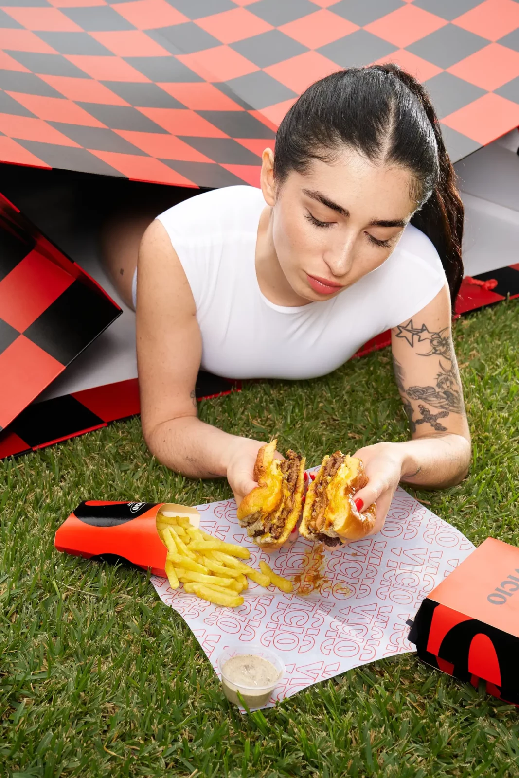 Overhead portrait of a tattooed model in a white tank top holding a burger over a patch of artificial grass.