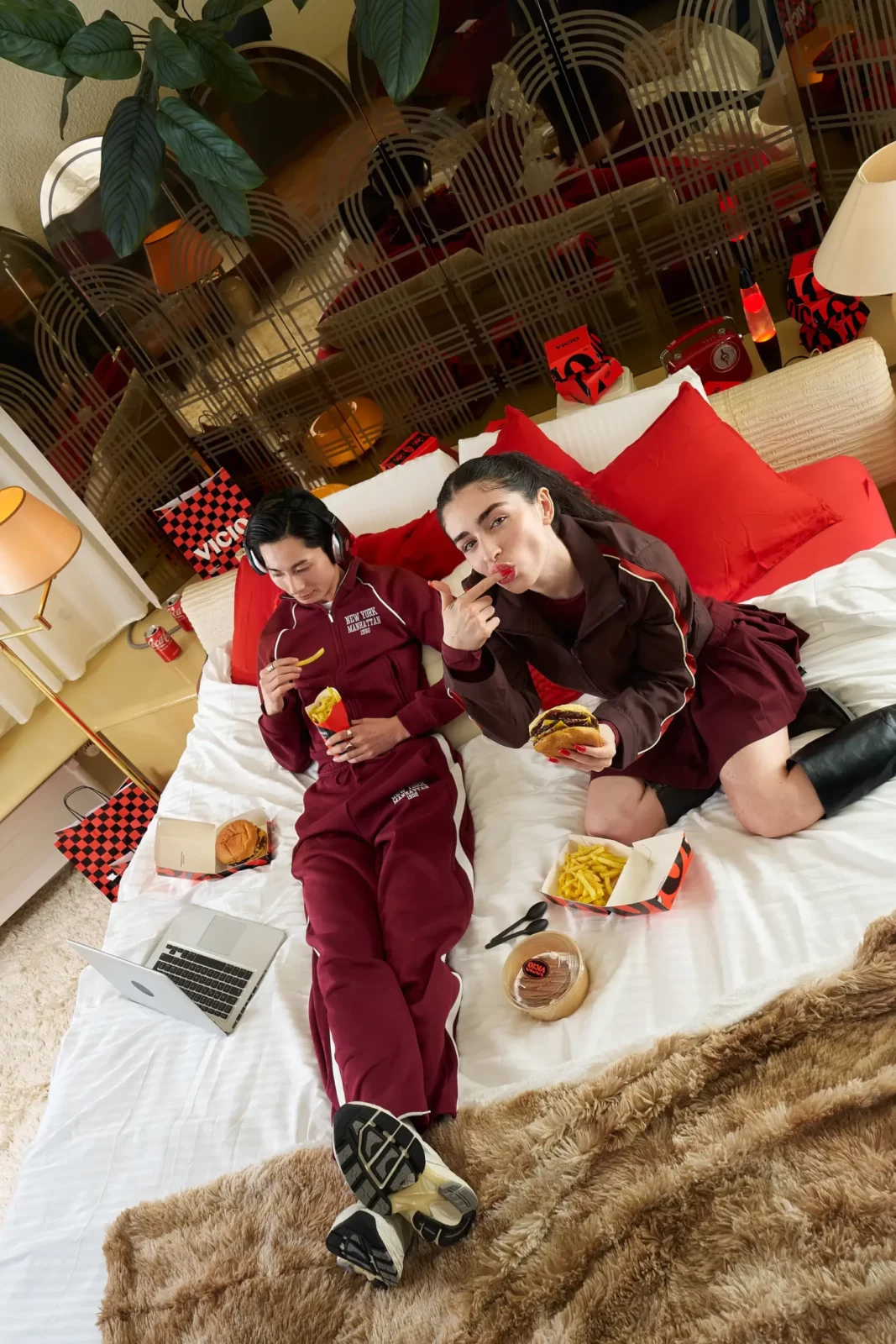 Wide overhead photograph of a model in a burgundy tracksuit reclining among food packaging and red cushions.