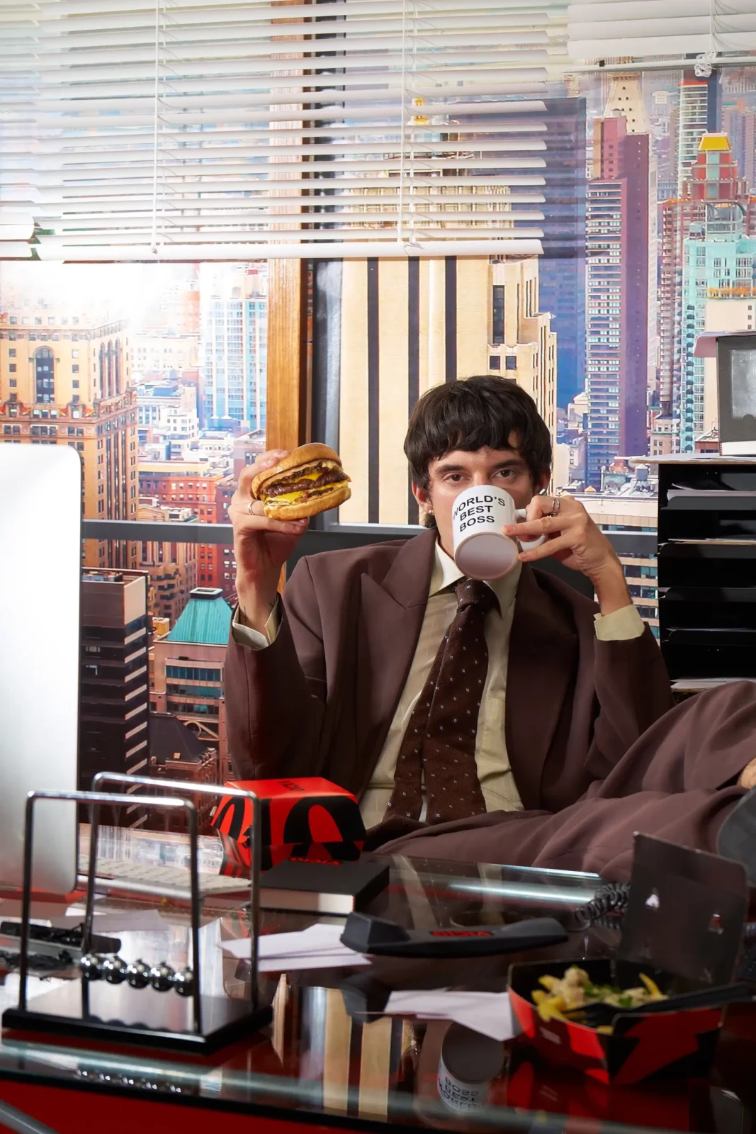 Fashion model in a brown suit and white shirt eating at a table near bright windows and city buildings.