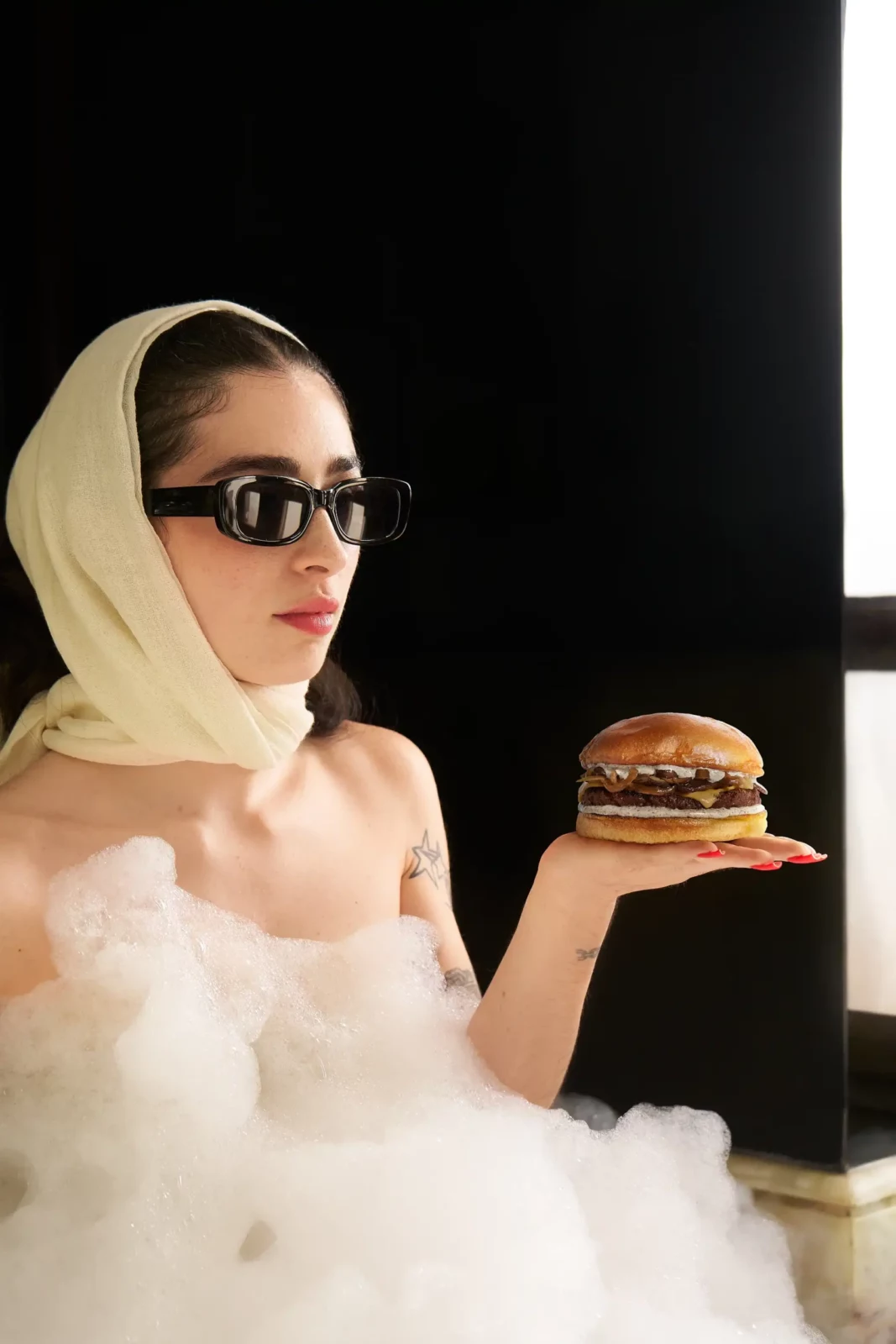 Blonde model in a sheer white outfit and dark sunglasses holding a hamburger against a black background.