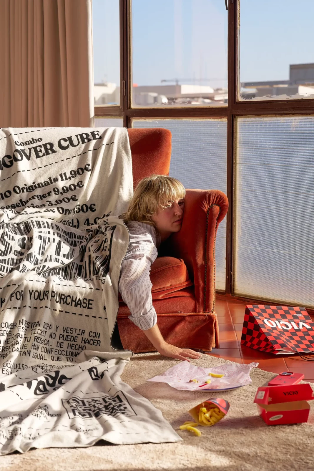 Blonde woman seated by large windows with food packaging and newspapers spread across a red room.