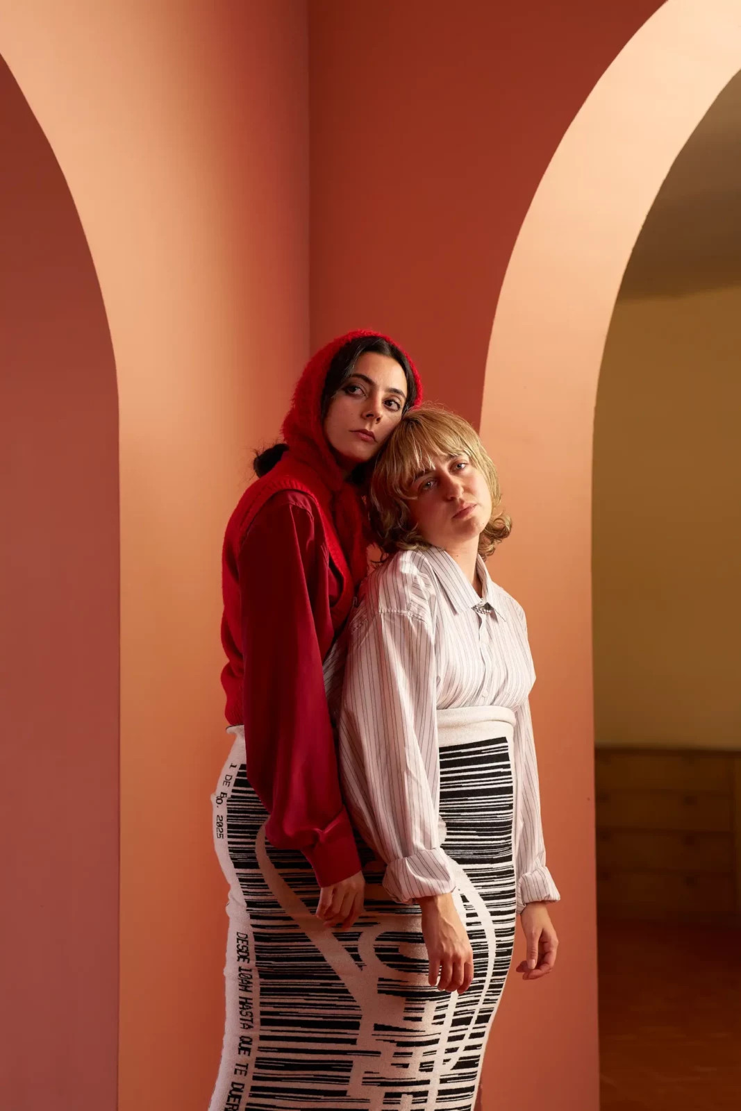 Two women wearing red and pale clothes standing beneath peach-colored arches with a patterned food box.