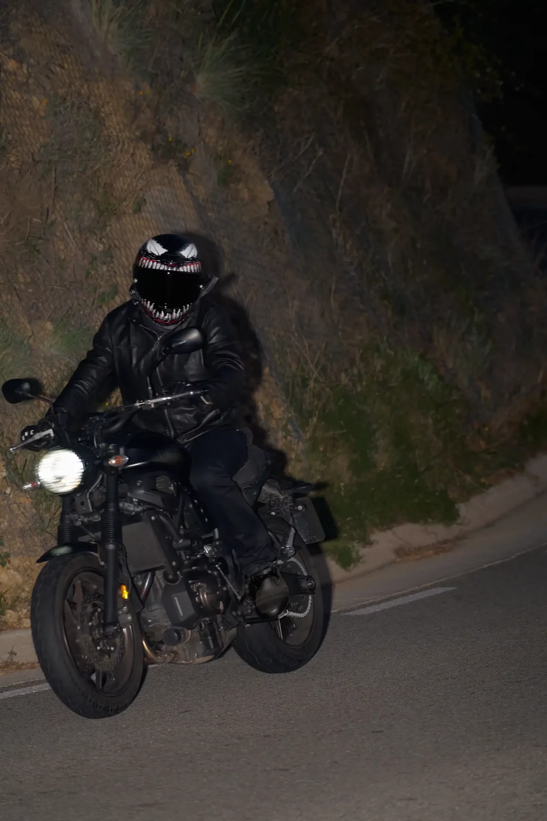 Full-length night photograph of a black-clad motorcyclist riding along a rough urban road.