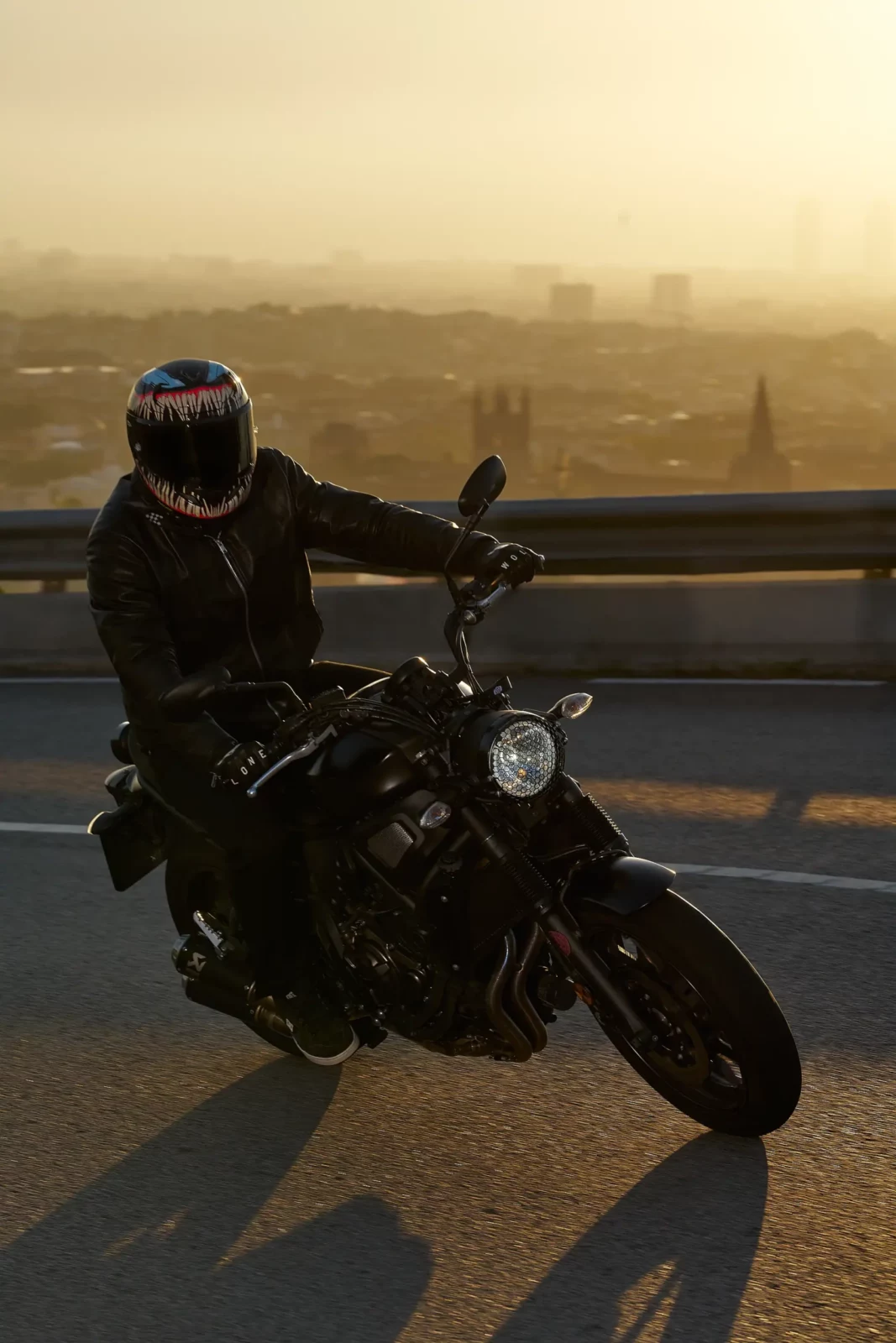Motorcyclist wearing a black helmet and jacket leaning on a motorcycle at sunset.