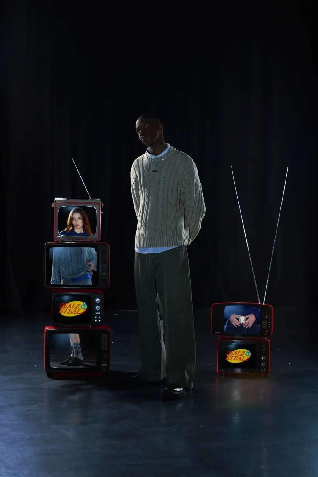 Standing model in an oversized pale outfit surrounded by glowing vintage televisions.