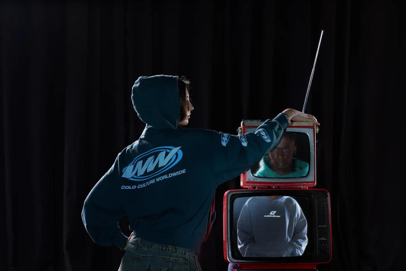 Rear view of a model in a blue hooded top touching a vintage television in a dark studio.
