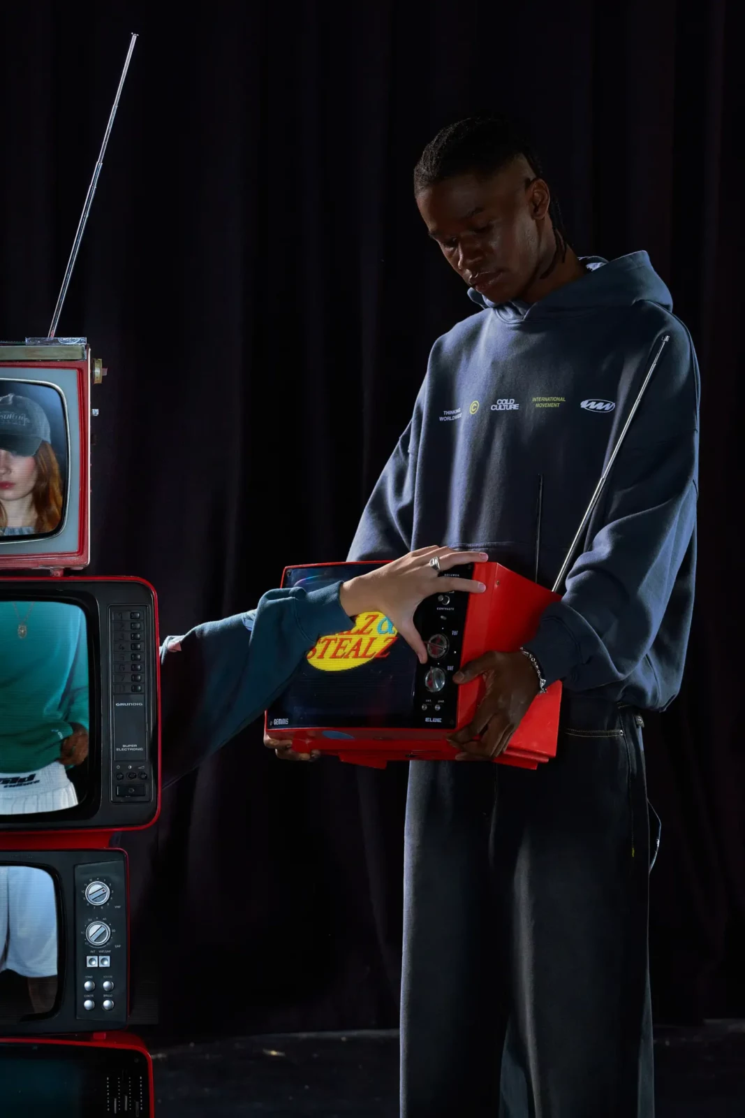 Person in a blue streetwear top holding a colorful garment beside stacked television screens.