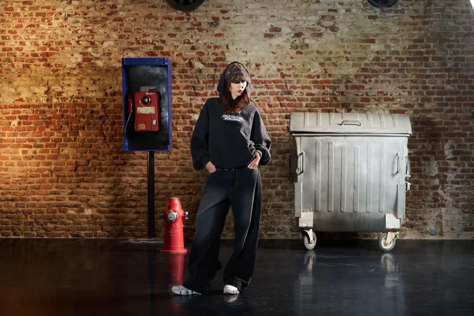 Streetwear model posing beside a vintage television and industrial metal bin against a brick wall.